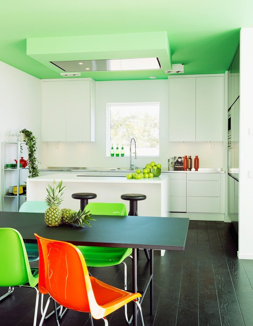 Colourful plastic chairs at black table, dark wooden floor, white modern counter and fitted kitchen in background below green-painted ceiling in open-plan interior