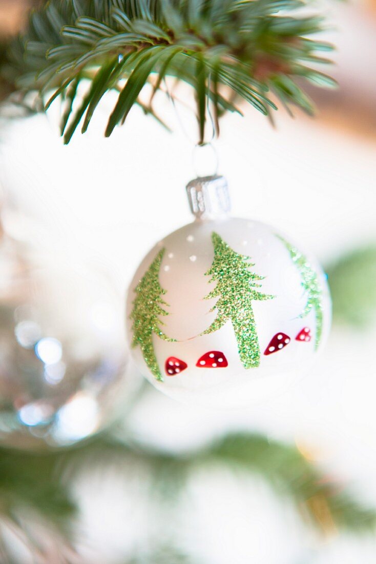 weiße Weihnachtskugel mit Flitteraufdruck stilisierter Tannenbäume und Fliegenpilze