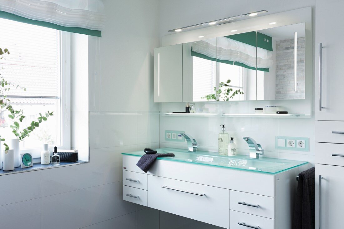 A double wash basin with a turquoise glass platter over a white cupboard with an illuminated, four-door mirrored cabinet