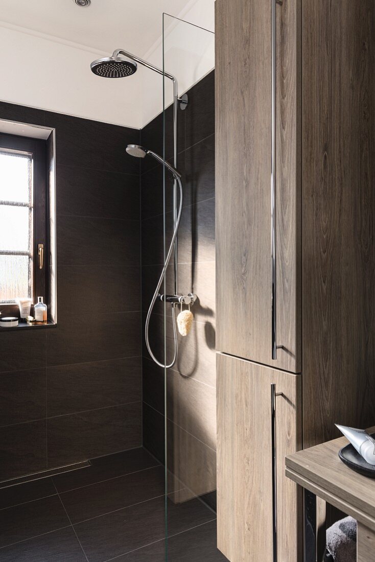 Bodenebene Dusche mit feststehender Glas Abtrennung, Kopf- und Handbrause an gefliester Wand, im Vordergrund Hochschrank mit Holzdekor Eiche und Edelstahl Griffleisten