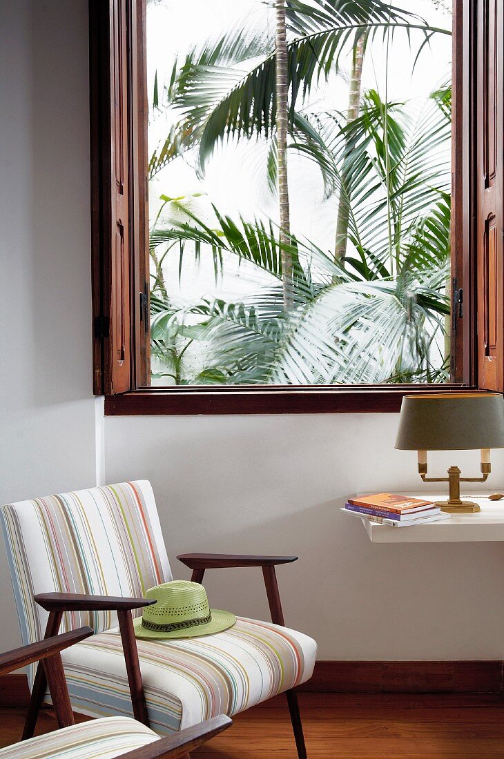 Sessel im 60er Jahre Stil mit Streifenbezug neben offenem Fenster und Blick auf Palmen