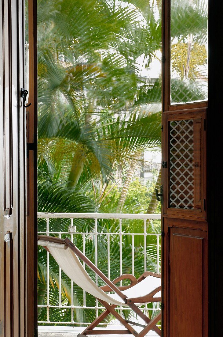 Blick durch offene Balkontüren auf Liegestuhl vor Palmen im Hinterhof