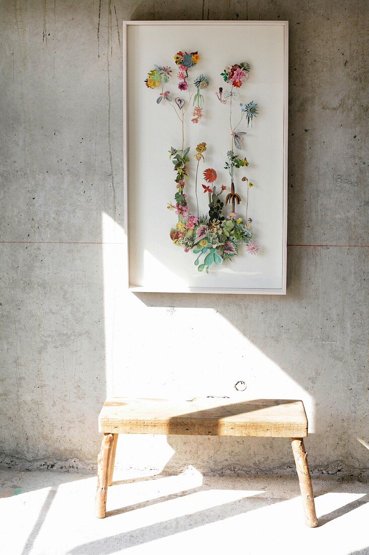 Schaukasten mit verschiedenen Blumen an Sichtbetonwand aufgehängt, davor rustikaler Holzschemel auf sonnenbeschienenem Boden