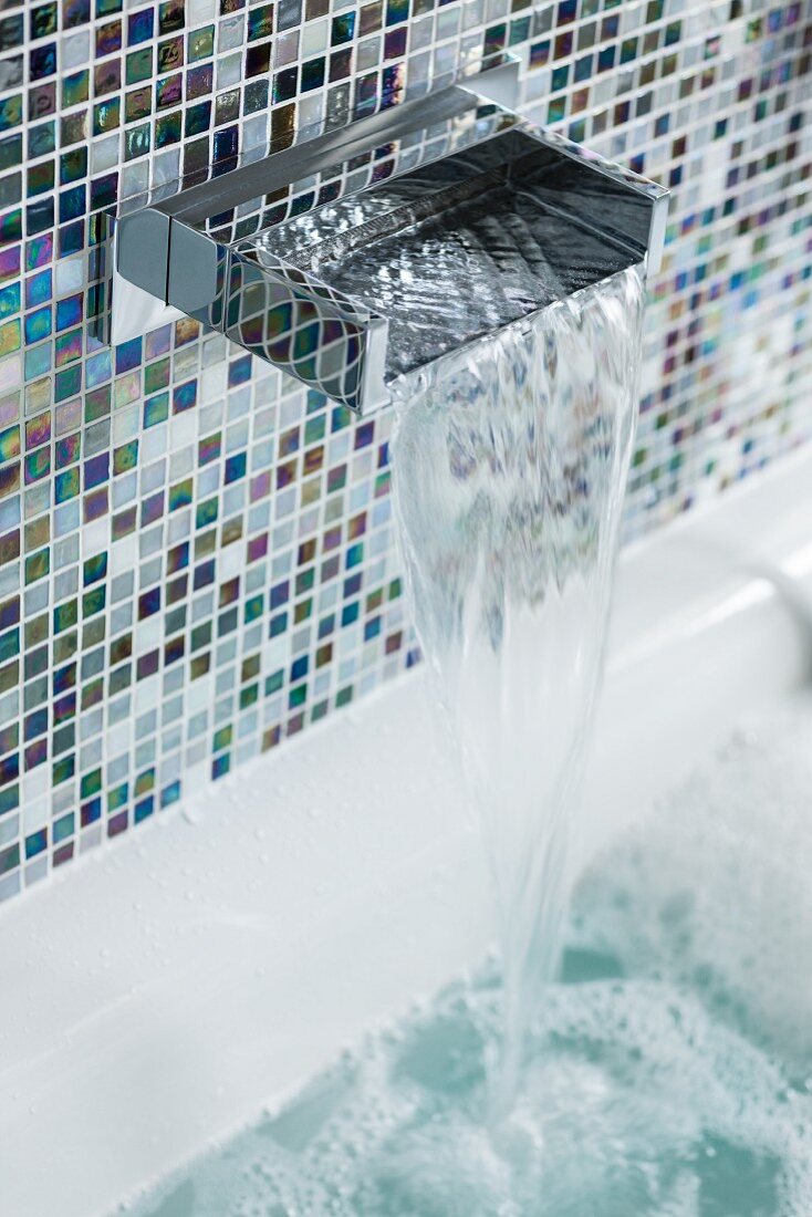 An elegant waterfall tap on a wall with shimmering blue-and-white mosaic tiles with the water running into a bubble bath