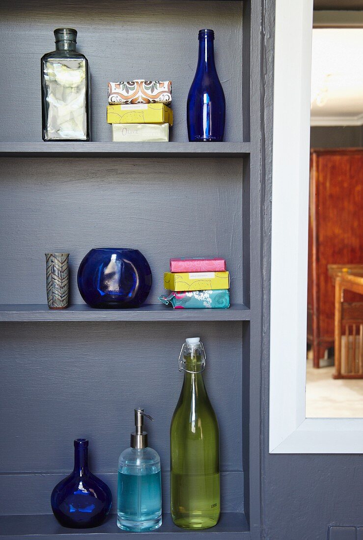 Various coloured glass bottles and brightly wrapped soaps on grey-painted shelves in niche