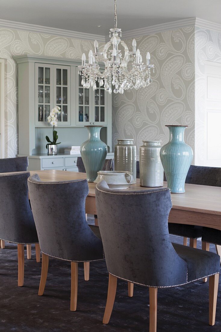 Chairs with blue velvet upholstery at long, pale wood dining table with collection of vases below crystal chandelier in traditional interior