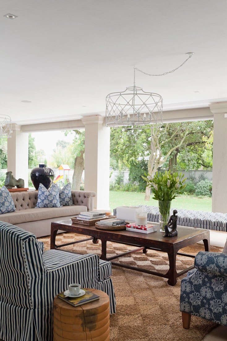 Sessel mit Streifenhusse und Couch um rustikalen Couchtisch aus dunklem Holz, im Hintergrund raumhohe Fenster mit Gartenblick