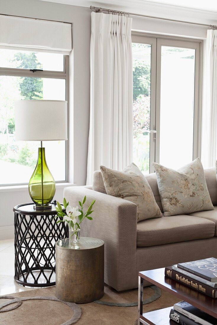 Verschieden hohe Beistelltische mit Blumenvase und Tischleuchte neben naturfarbenem Sofa, im Hintergrund weisser Vorhang an Terrassentür in traditionellem Ambiente