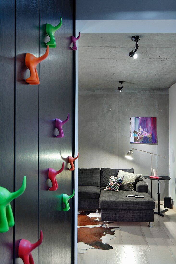 Colourful plastic hooks on black wall next to open doorway leading to living room with grey corner sofa against concrete wall
