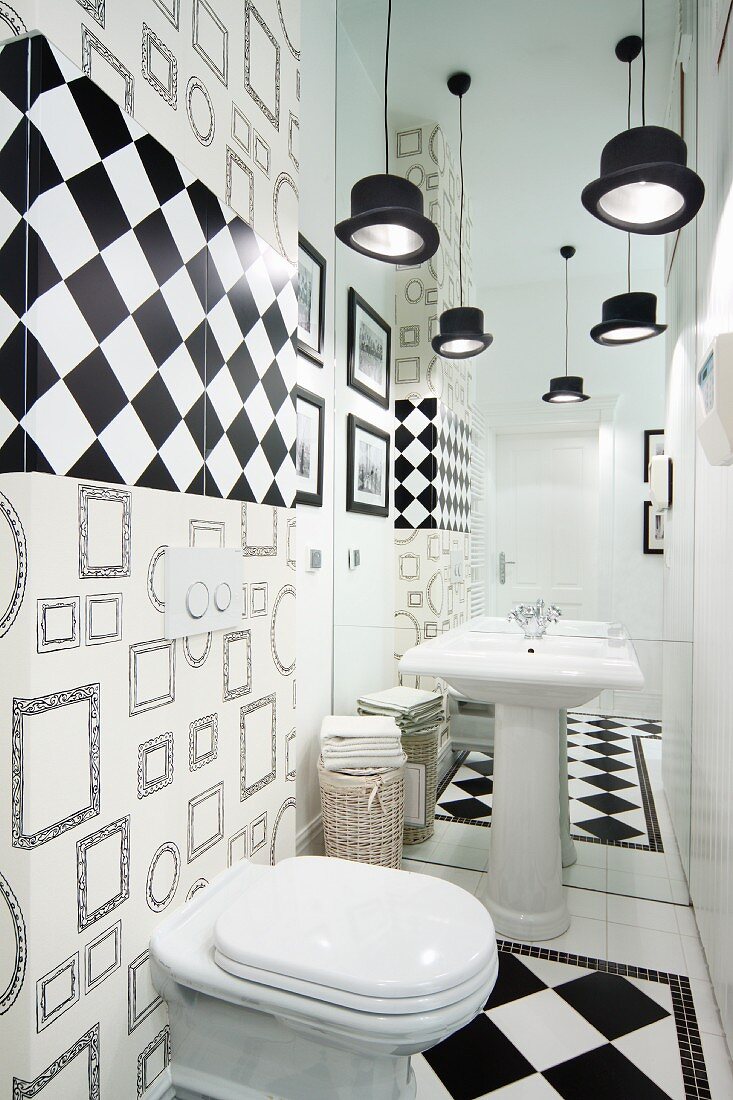 Toilet decorated in black and white with top-hat pendant lamps and and illusion of space produced by mirrored wall behind pedestal sink
