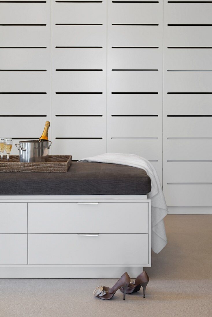 Ladies' shoes and half-height chest of drawers with upholstered top in front of white fitted cupboards with slotted doors