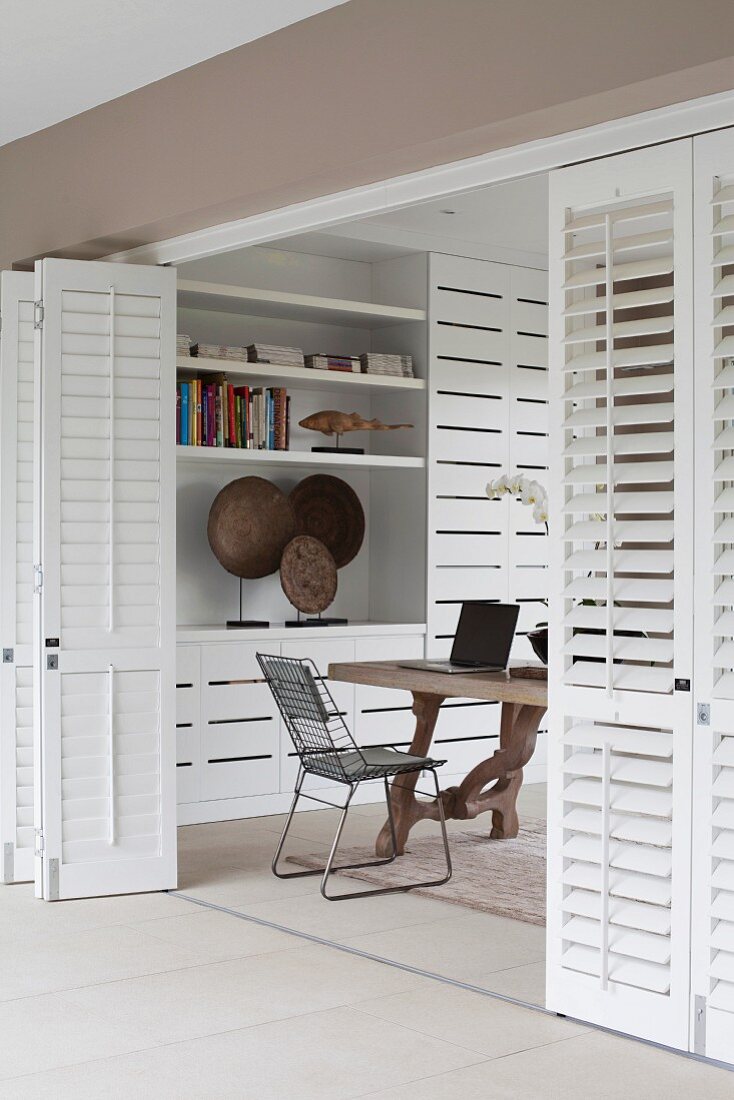 Partially open, white folding doors with view of chair at rustic wooden table and modern fitted cupboards