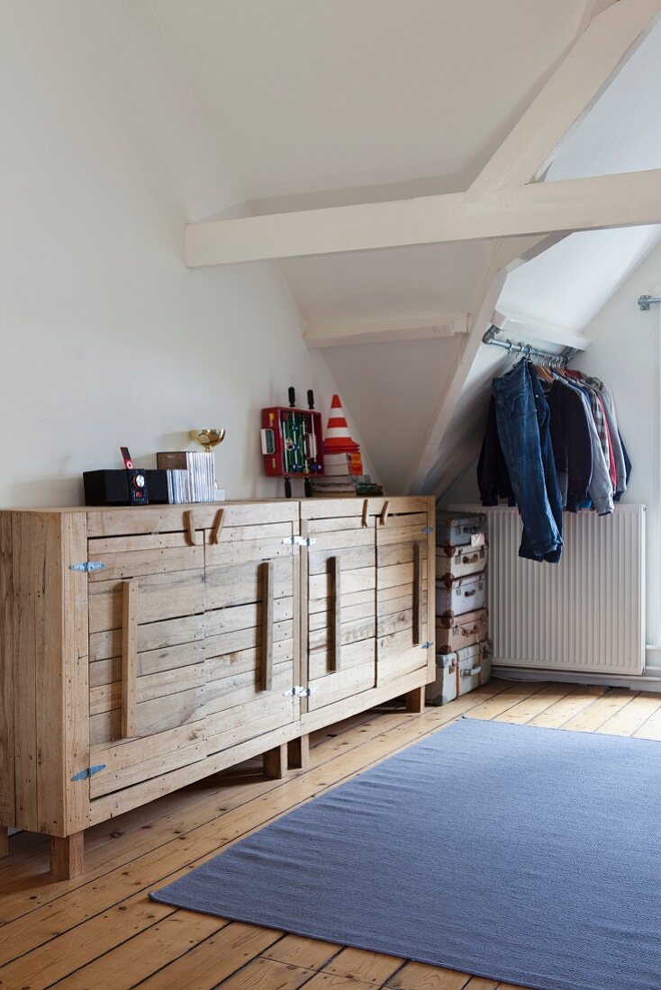 Rustic, plain wood sideboard in attic room; clothing hung from metal rod and stacked suitcases in background
