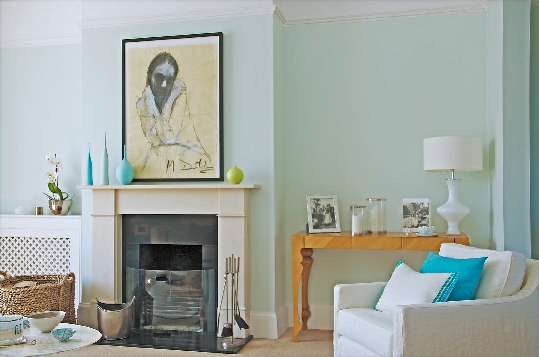 Open fireplace and white armchair in front of family photos on artistic console table against pastel wall in elegant interior