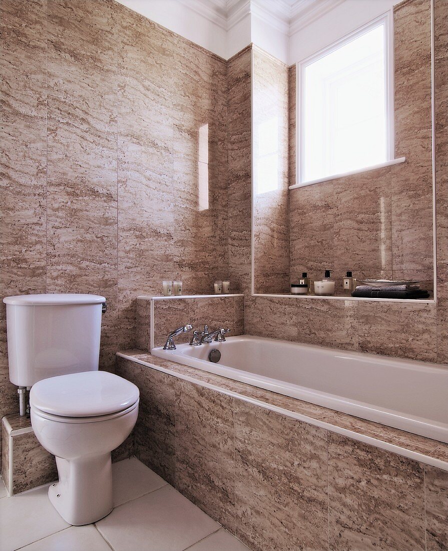 Masculine atmosphere in simple, beige, marble bathroom with tiled walls, bathtub below window and traditional, white toilet with cistern