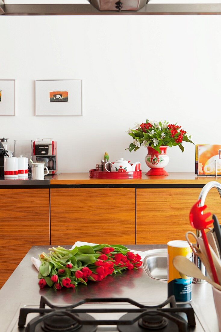 Ausschnitt einer Kochinsel, rote Tulpen auf Edelstahl Arbeitsplatte, gegenüber Sideboard aus hellem Holz