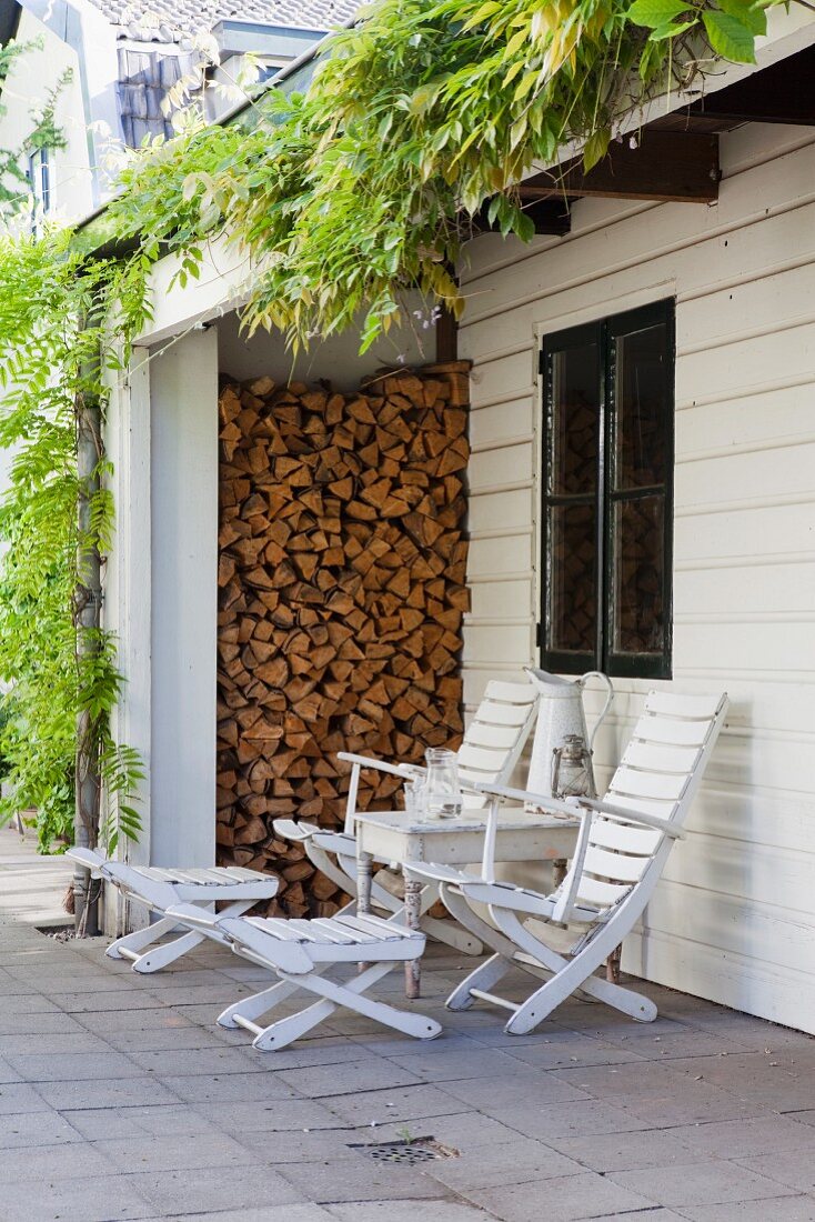 Seating area with white deckchairs and side table below wisteria growing over outside one-storey extension with stacked firewood