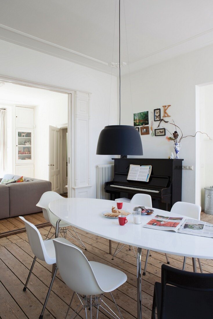 Schwarze Pendelleuchte über weißem Ovaltisch mit Schalenstühlen; im Hintergrund ein Klavier und das Sofa im angrenzenden Wohnraum
