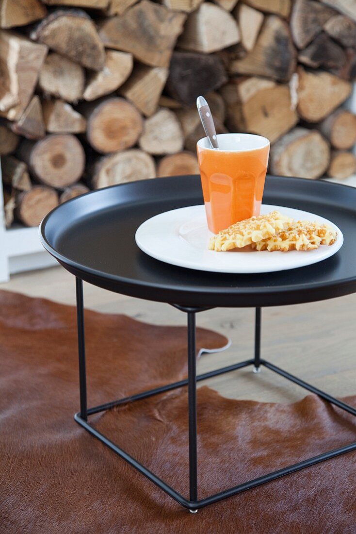Beaker of coffee and waffles on charcoal tray table on animal skin rug in front of stacked firewood