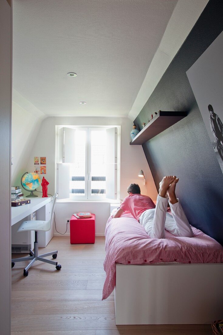 Auf dem Bett liegender Jugendlicher, gegenüber eingebauter weißer Schreibtisch im schmalen Dachzimmer