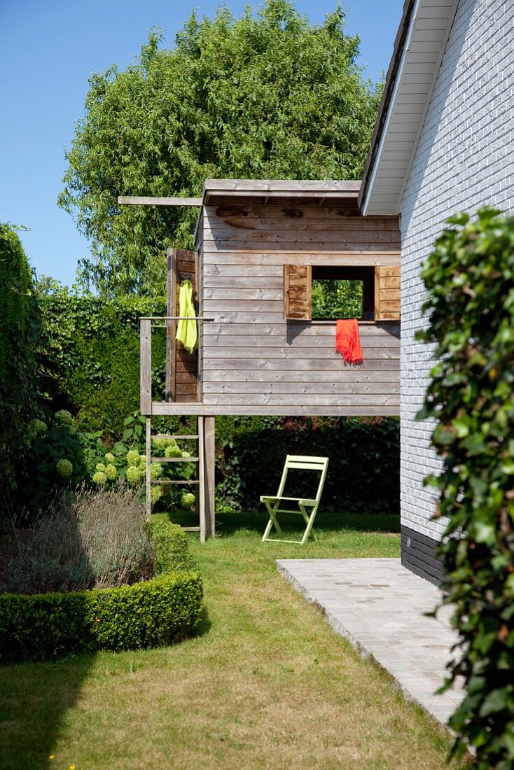 Wooden playhouse on stilts in garden surrounded by tall hedge; whitewashed brick facade to one side