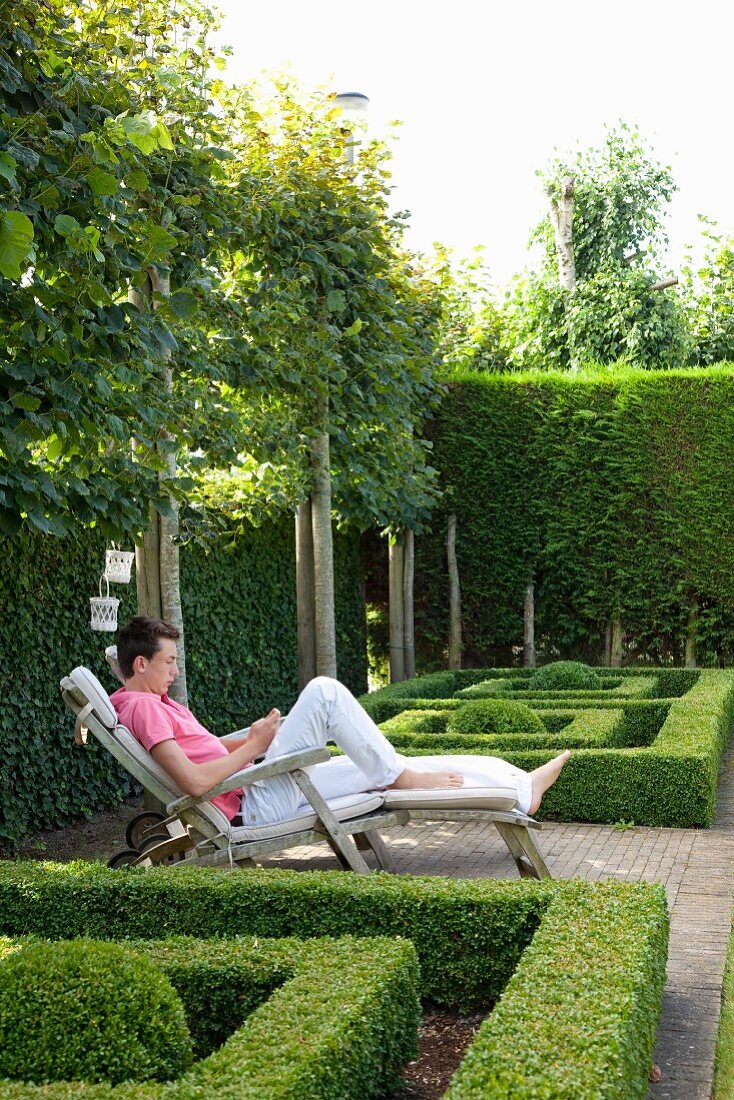 Boy sitting on deckchair amongst geometric box hedges in garden surrounded by espalier trees and tall hedges