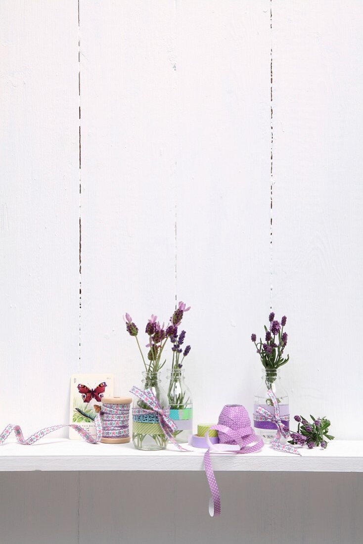 Sprigs of lavender in glass bottles decorated with ribbons and washi tape on surface against whitewashed wooden wall