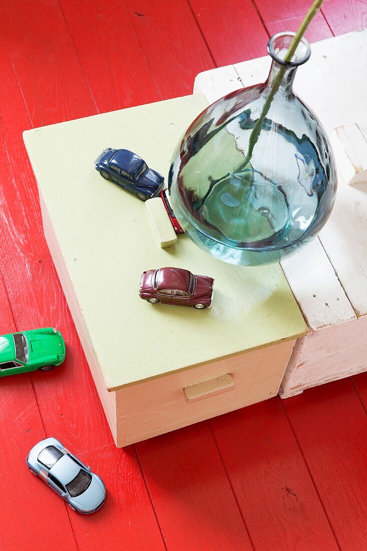 Glasvase und Spielzeugautos auf Holzschachteln und rot lackiertem Holzboden