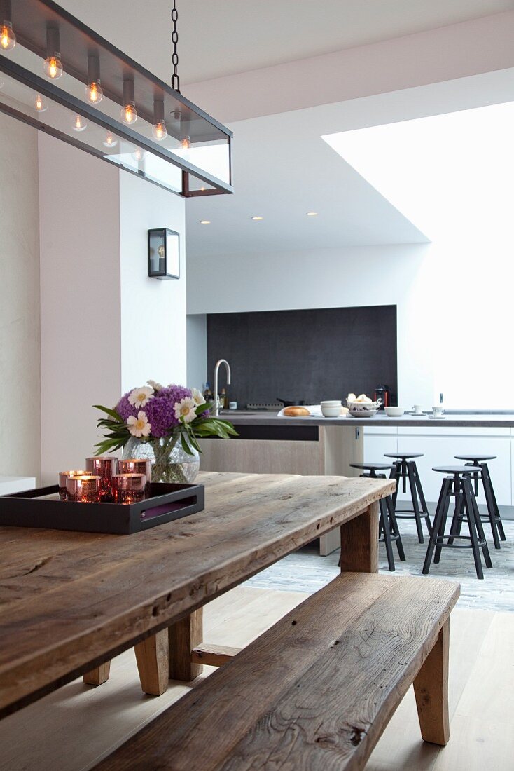 Modern, wrought iron lamp above rustic dining area with wooden table and wooden bench; stools at kitchen counter in background