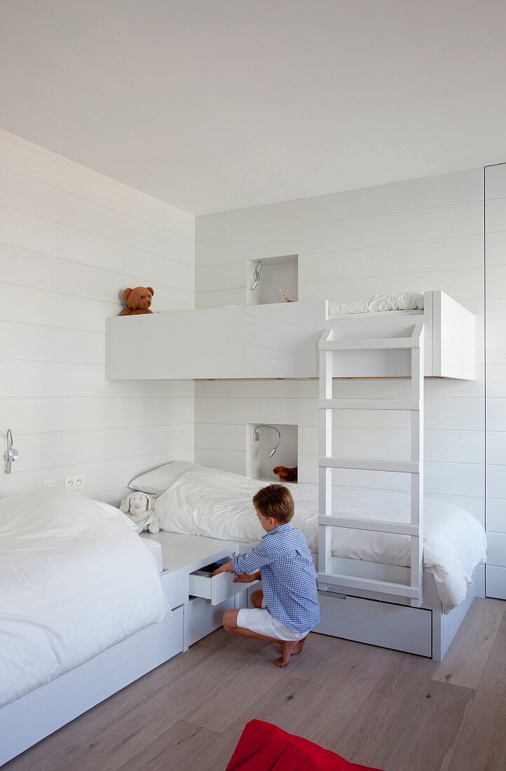 Two beds arranged in an L and one loft bed in children's bedroom painted white; boy opening drawer in base