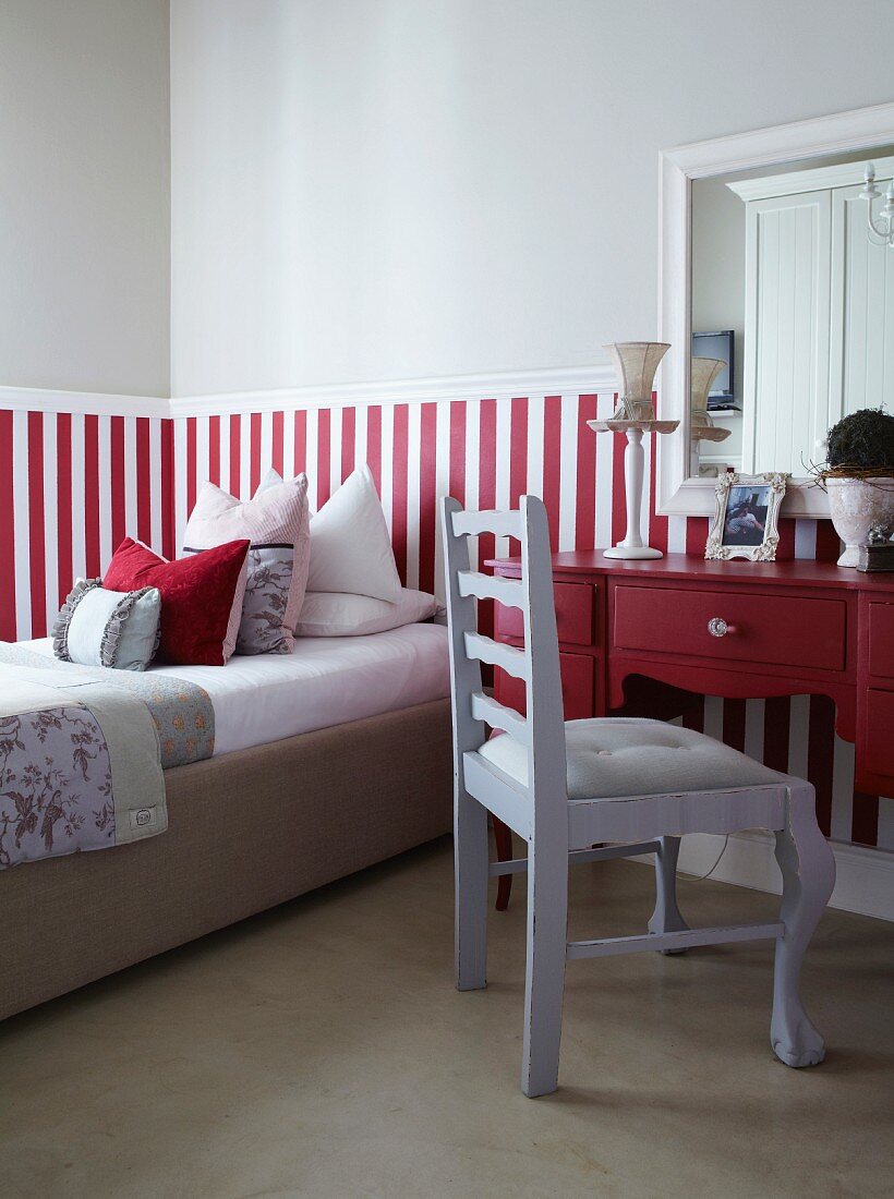 Hellgrau lackierter Stuhl vor rotem Schreibtisch, daneben Bett vor halbhoch, rotweiss gestreifter Wand