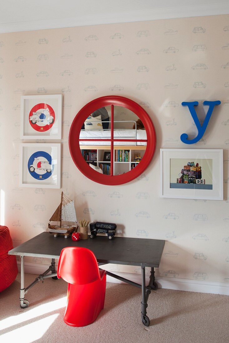 Roter Kinderstuhl aus Kunststoff vor mobilem Tisch, an tapezierter Wand runder Spiegel mit rotem Rahmen zwischen gerahmten Bildern