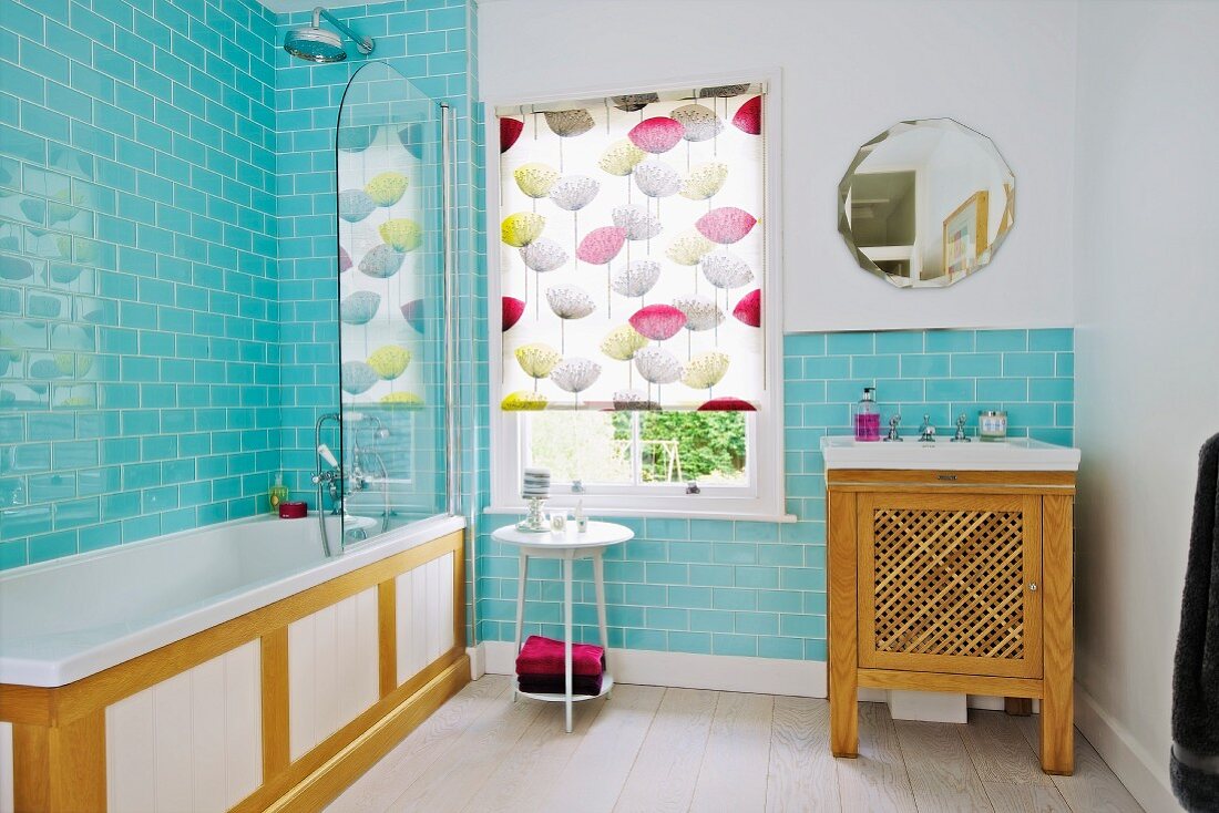 Bathroom with pale blue wall tiles, wood-clad bathtub, patterned roller blind on window and sink on wooden washstand