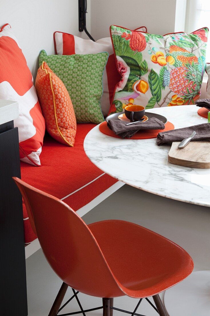 Orangefarbener Klassikerstuhl vor Tisch mit Marmorplatte, seitlich drapierte Kissen auf Sitzbank