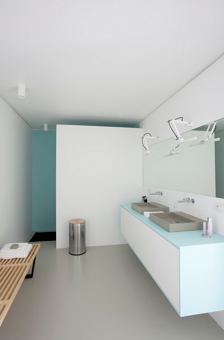 Designerbad mit weißem Waschtischmöbel und zwei Waschbecken, langer Spiegel mit Wandbeleuchtung, im Hintergrund weiße Wand vor Duschbereich