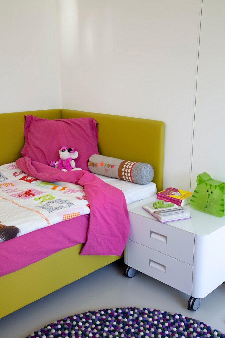 Comfortable bed with lime green upholstered frame and white cabinet on castors as bedside table in child's bedroom