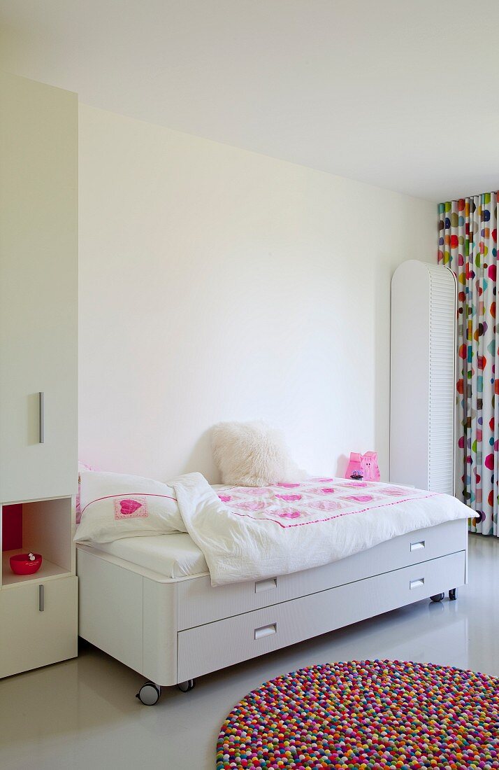 Round, colourful rug in front of white bed on castors with storage drawer in bright, girl's bedroom