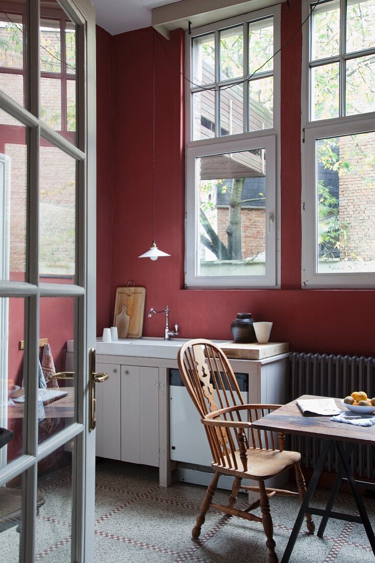 Blick durch offene Sprossentür auf Armlehnstuhl mit hoher Rückenlehne am Esstisch, in schlichter Küche mit bordeauxrot getönter Wand
