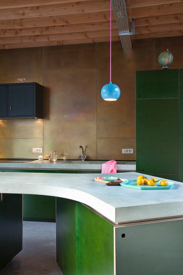 Spherical, blue lamp above curved island counter with green base units; back wall of kitchen clad in brass panels