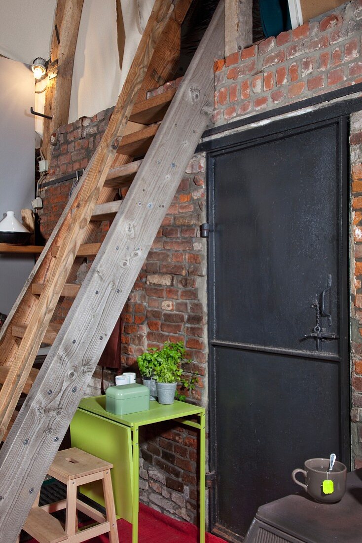 Wooden ladder leading to gallery level in loft-apartment kitchen; brick wall with industrial, black metal door