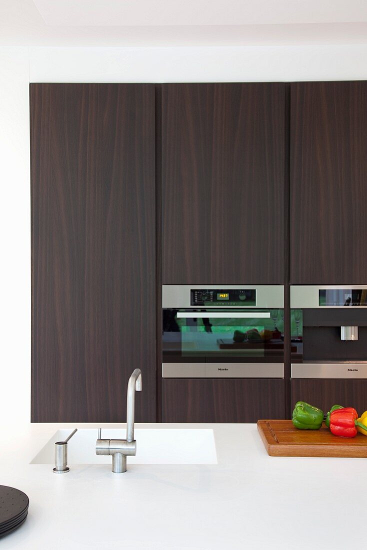 Blick über Kücheninsel mit Corian-Arbeitsfläche auf dunkle Einbaufronten und Einbaugeräte einer Designerküche
