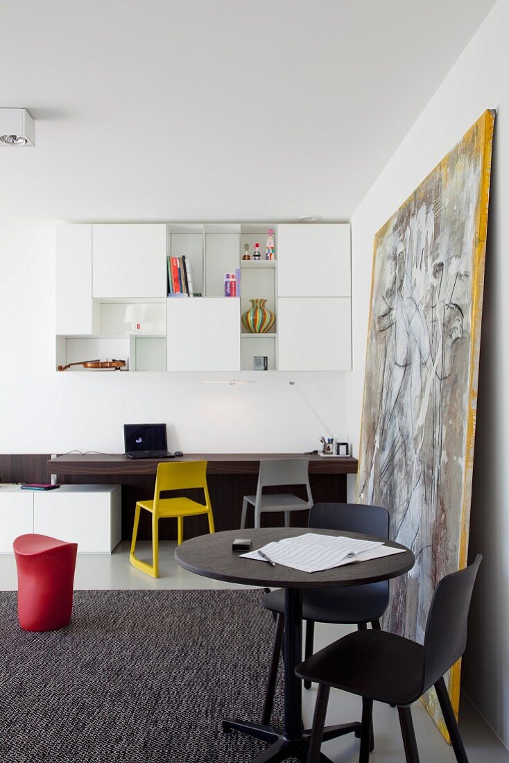 Round table and black chairs, desk and colourful chairs and white, wall-mounted cabinets in modern interior