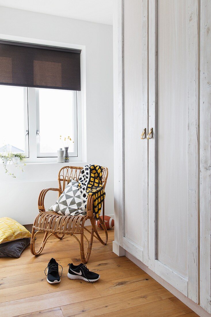 Retro-style cane armchair in corner below window next to white-painted wooden wardrobes