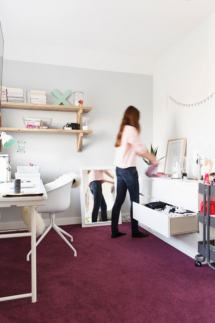 Weisser Drehstuhl am Schreibtisch, gegenüber Mädchen beim Einräumen vor offener Kommode im Jugendzimmer mit violettem Teppich