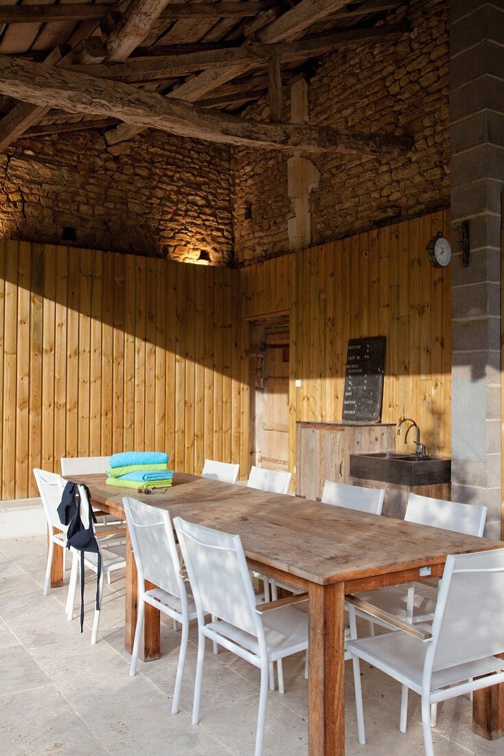Outdoor-Küche - Lange Holztafel und weiße Metallstühle auf rustikaler, überdachter Terrasse mit Holzverschalung