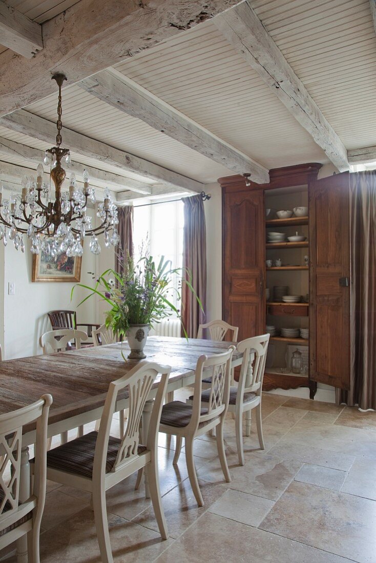 Weiß lackierte Küchenstühle mit geschnitzter Rückenlehne um Holztisch, im Hintergrund Geschirrschrank in rustikalem Esszimmer mit Holzbalkendecke