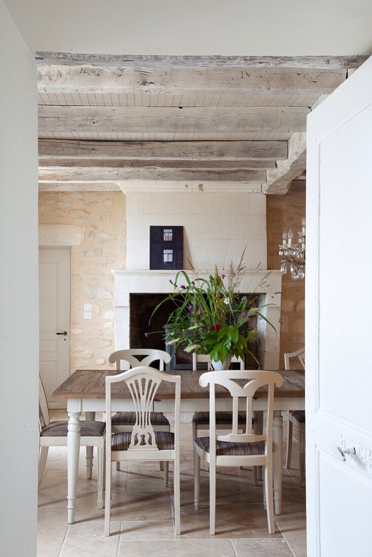 White-painted kitchen chairs around wooden table in front of fireplace in rustic dining room