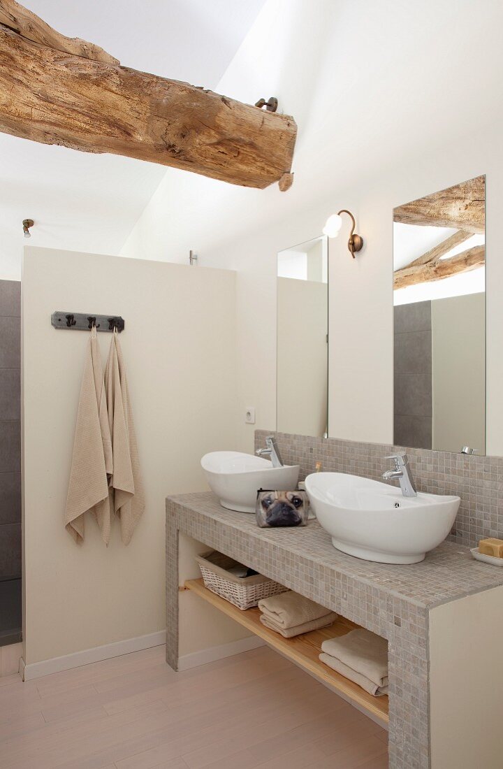 Masonry washstand covered in mosaic tiles and white countertop basins below mirror on wall in renovated bathroom with wooden ceiling beam