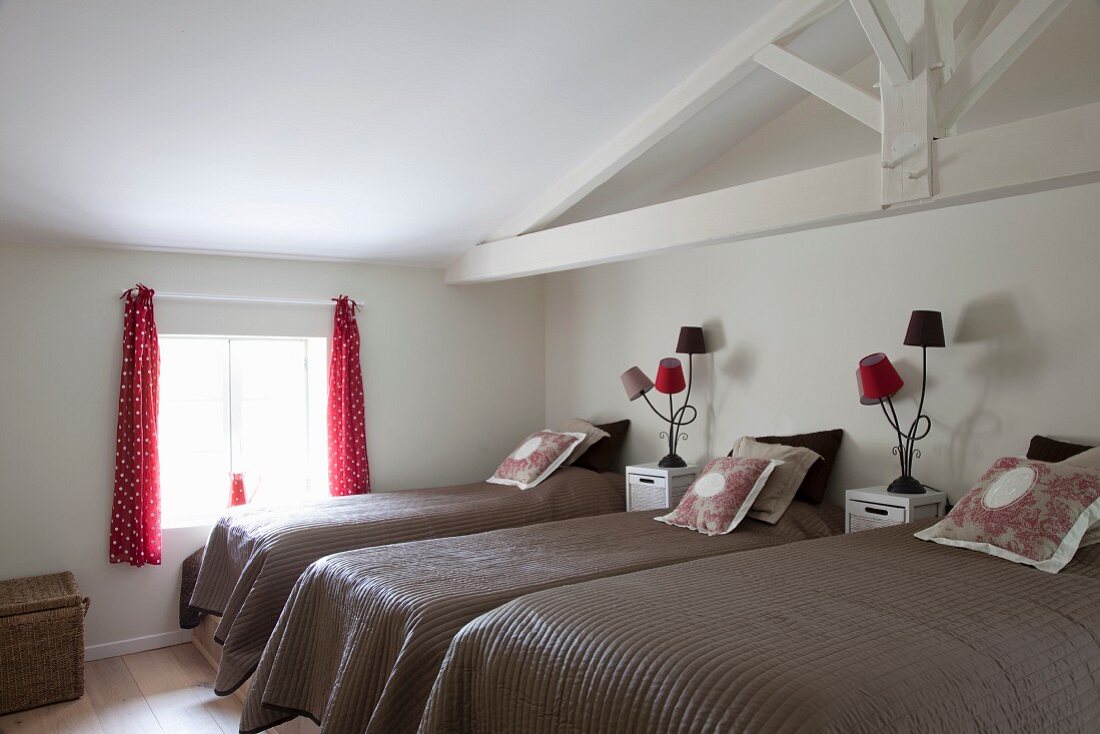 Three single beds with brown bedspreads, bedside cabinets and table lamps with small, red lampshades in attic room with white, exposed roof structure