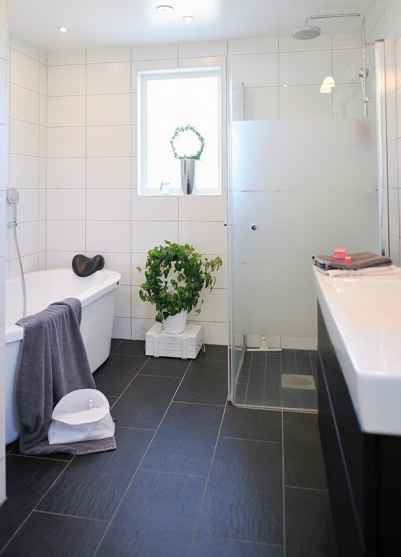 Weisses Badezimmer mit verglaster Duschkabine und dunkler Fliesenboden, im Hintergrund Zimmerpflanze unter Fenster